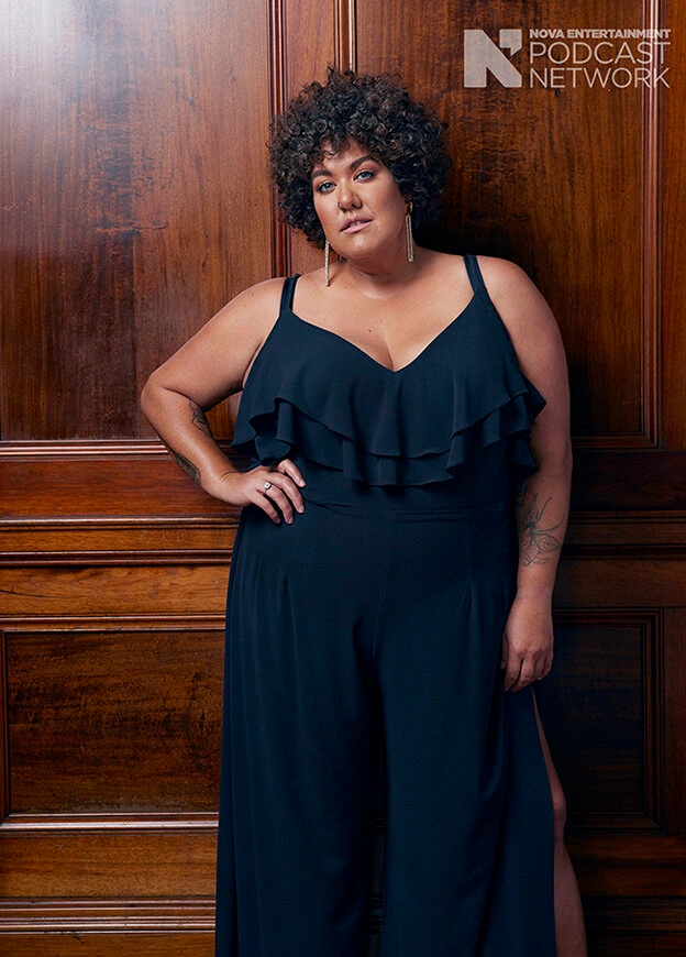 Woman with dark curly hair wearing a black jumpsuit posing against wood paneling with podcast network logo
