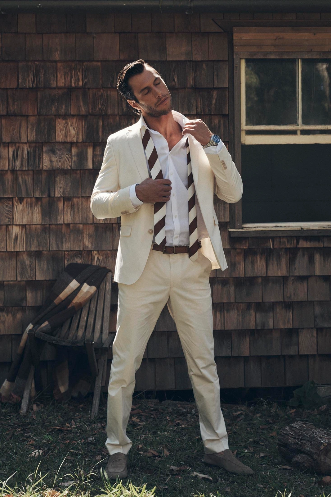 Man wearing a cream suit and white shirt adjusting a striped tie outdoors by a wooden building.