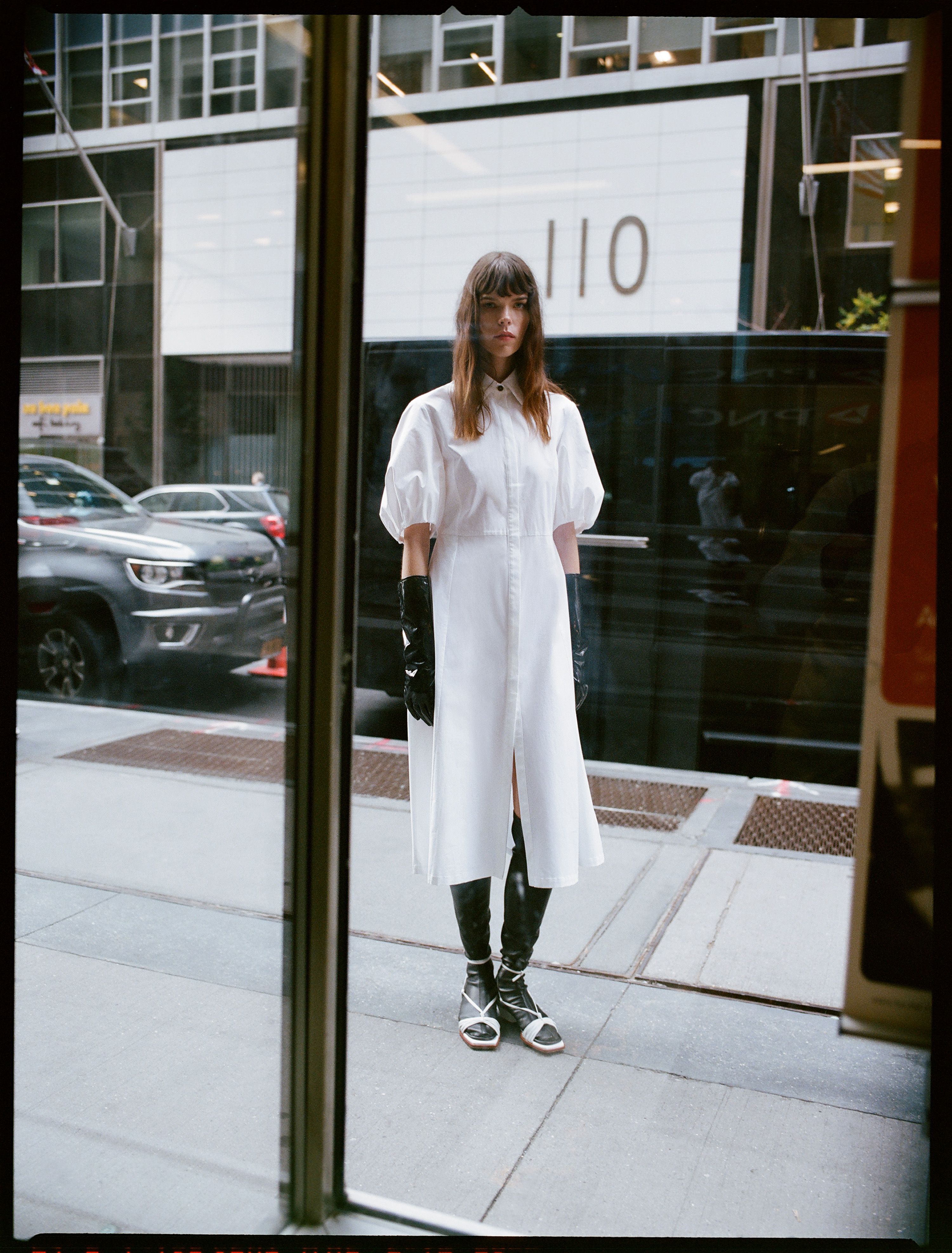 A woman stands in front of a building wearing a white dress, black gloves, and boots; a city street scene is visible through the glass.