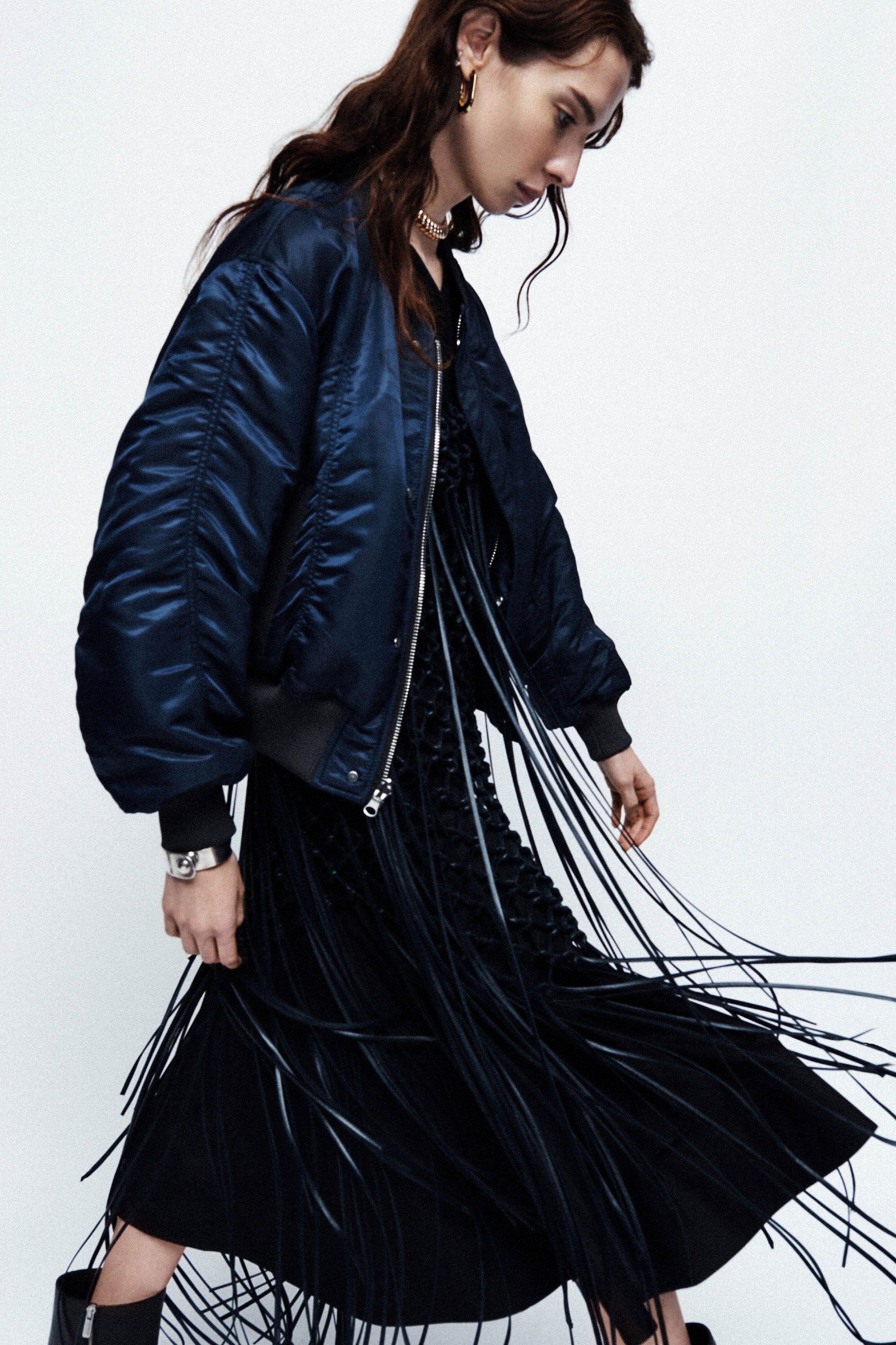 A fashionable woman in a blue bomber jacket and black dress with fringe poses against a white backdrop in a studio shot.