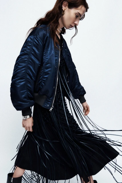 A fashionable woman in a blue bomber jacket and black dress with fringe poses against a white backdrop in a studio shot.