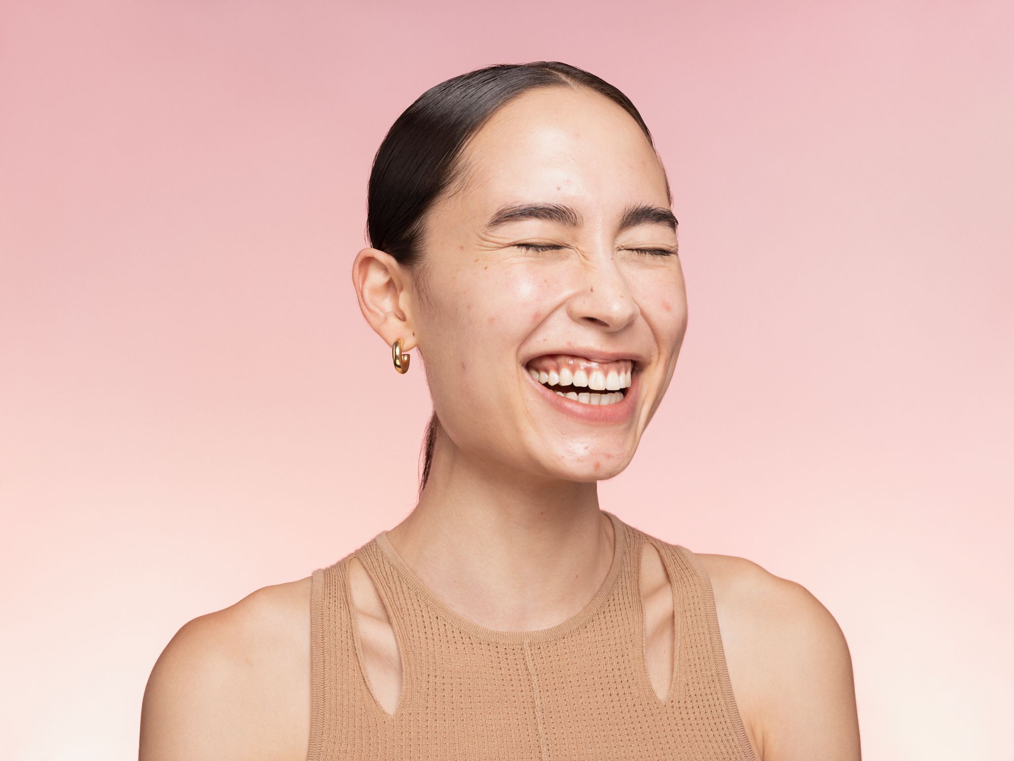 Portrait of a young woman with tan skin laughing with her eyes closed, wearing a tan top and gold hoop earrings against a pink gradient background.