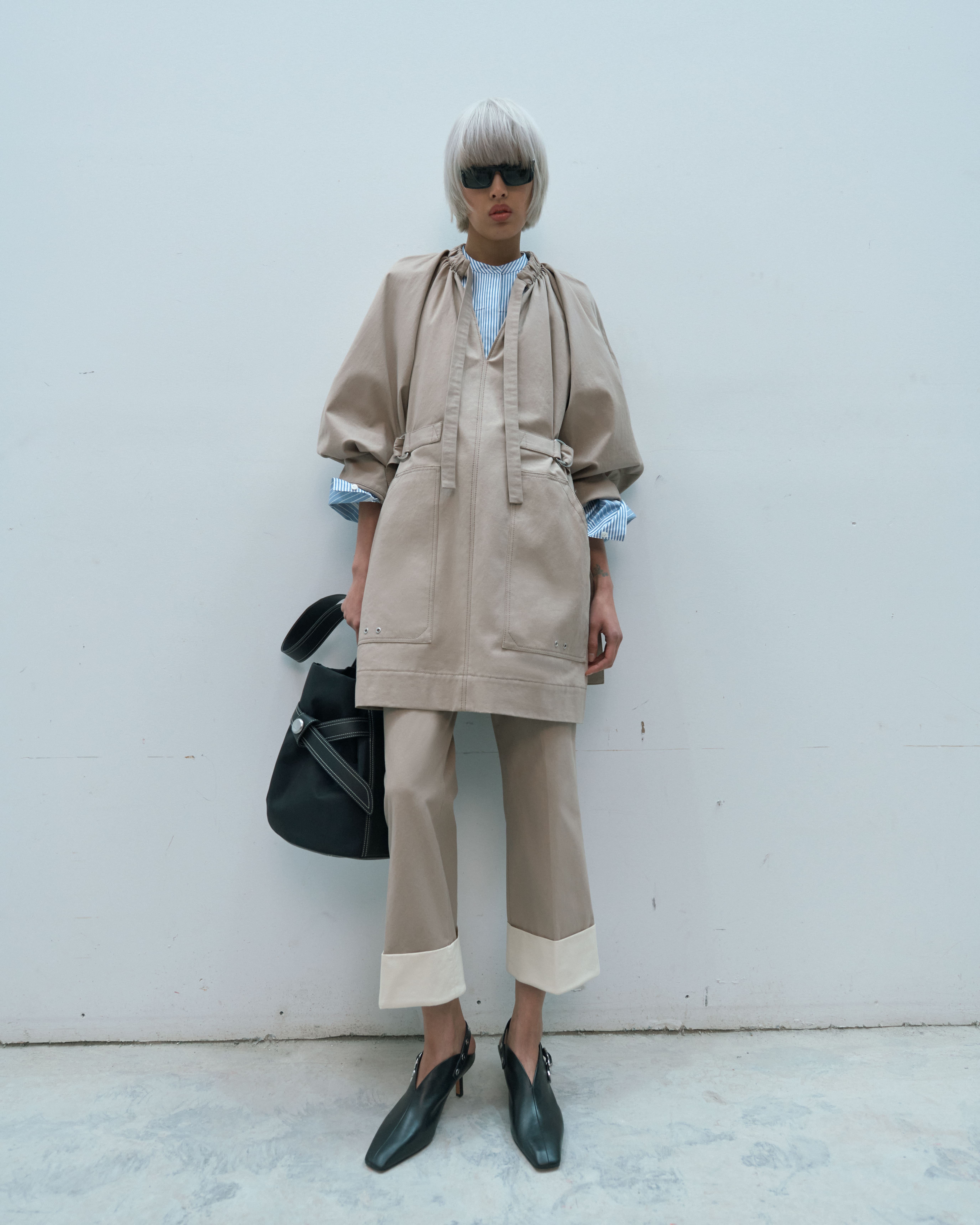Fashionable woman in a beige dress and pants poses against a white wall, wearing black sunglasses and carrying a black bag.