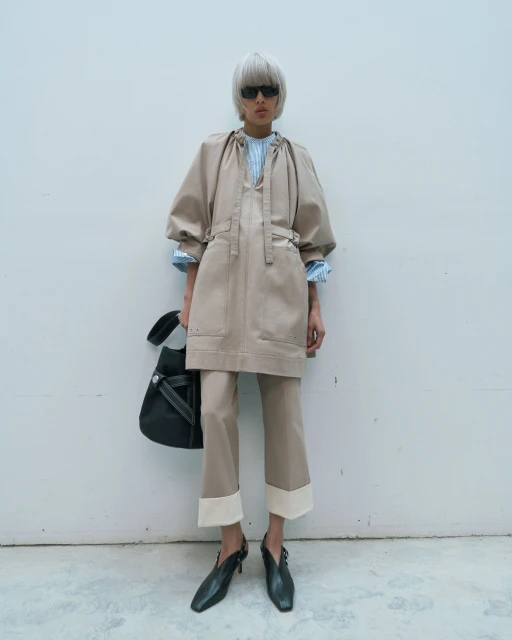 Fashionable woman in a beige dress and pants poses against a white wall, wearing black sunglasses and carrying a black bag.