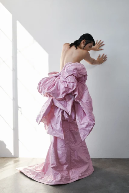 An adult leaning against a white wall wearing a couture pink gown constructed from crinkled vinyl, in a studio with directional lighting.