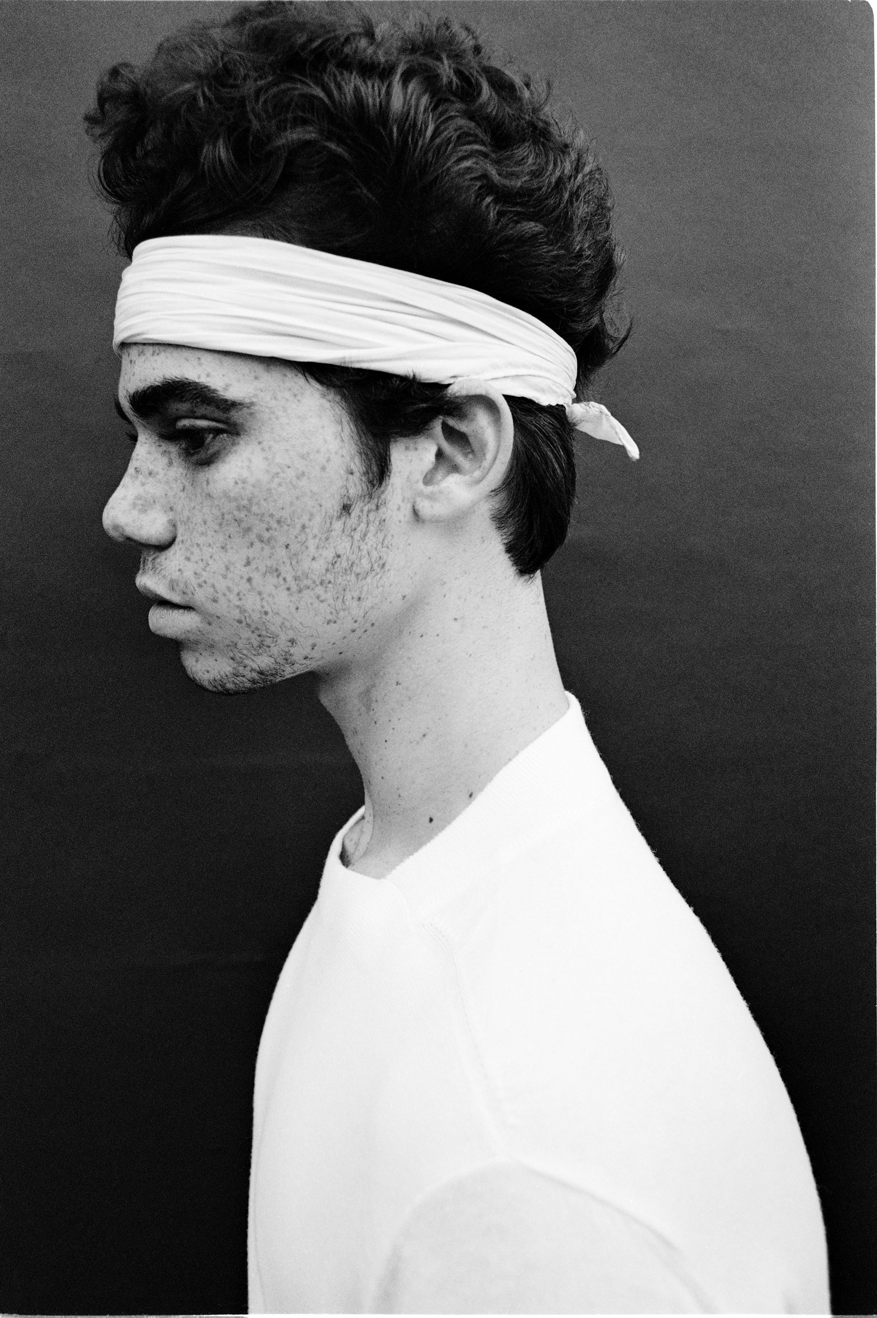 Monochrome portrait of a young adult male with curly hair and a white headband, captured in a studio setting against a dark background.