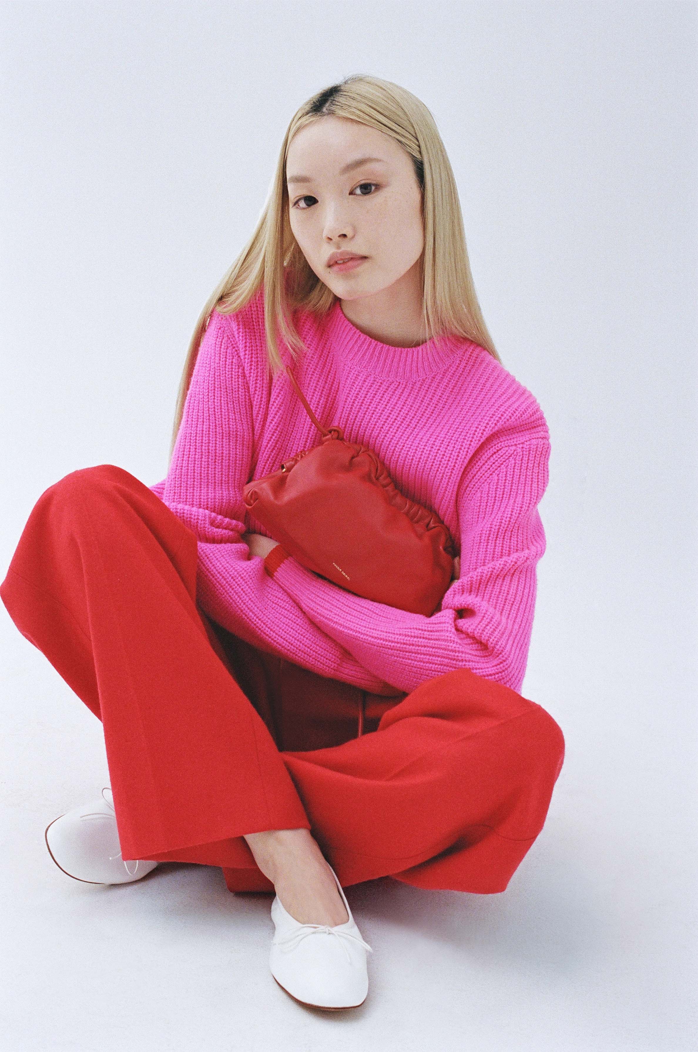 Asian female model poses cross-legged in a pink sweater, red pants, white flats and a red purse in a bright studio.