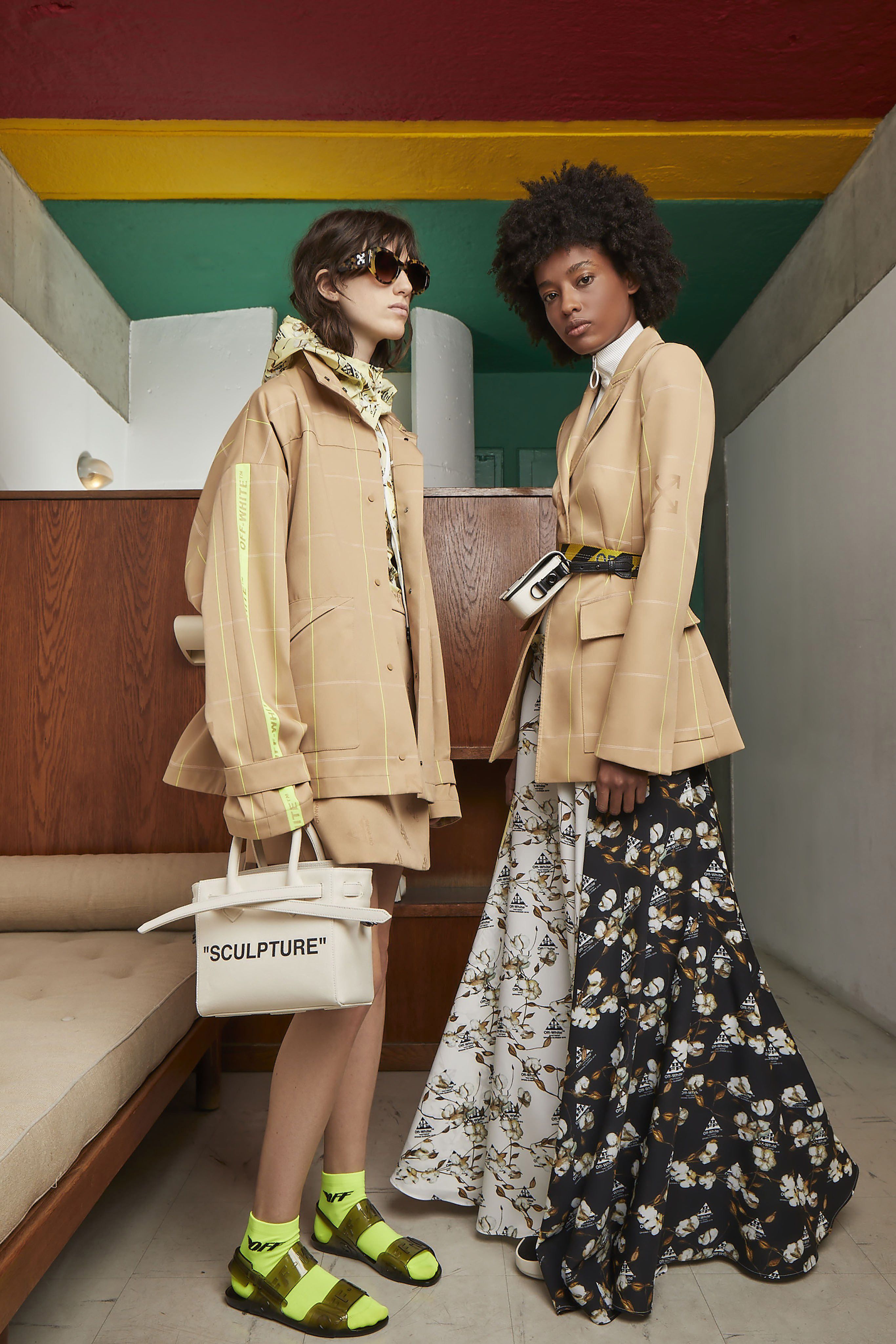 Two fashion models pose in jackets, skirts, and unique shoes; one carries a 'Sculpture' handbag in an indoor setting with colorful accents.