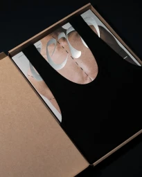 Top-down view of a black tank top placed over a magazine inside an open cardboard shipping box.