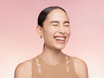 Close-up portrait of a laughing woman wearing a beige cutout top against a gradient pink backdrop, styled by Melissa Levy.