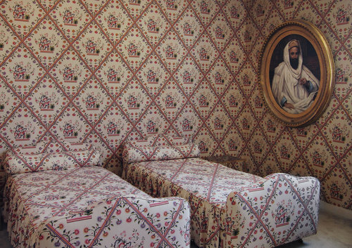 Vintage bedroom interior with two twin beds featuring floral patterned bedding and matching wallpaper, plus a framed portrait hanging above.