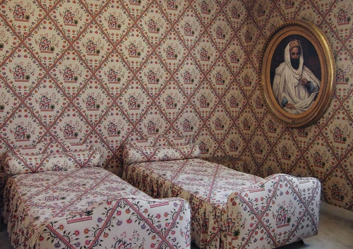 Vintage bedroom interior with two twin beds featuring floral patterned bedding and matching wallpaper, plus a framed portrait hanging above.