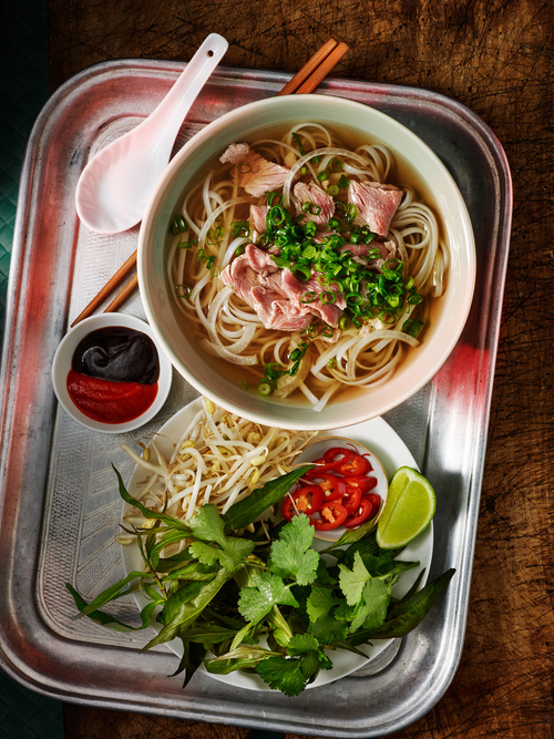 Overhead shot of Pho soup with noodles, sliced meat, and green onions in a bowl, with bean sprouts, herbs, lime, and sauces on a tray.