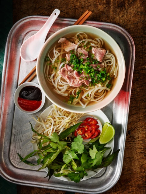 Overhead shot of Pho soup with noodles, sliced meat, and green onions in a bowl, with bean sprouts, herbs, lime, and sauces on a tray.