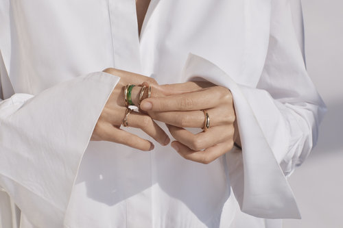 Close-up of a person wearing gold rings on their fingers, showcasing fashion jewelry against a white collared shirt and plain background.