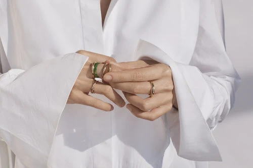 Close-up of a person wearing gold rings on their fingers, showcasing fashion jewelry against a white collared shirt and plain background.