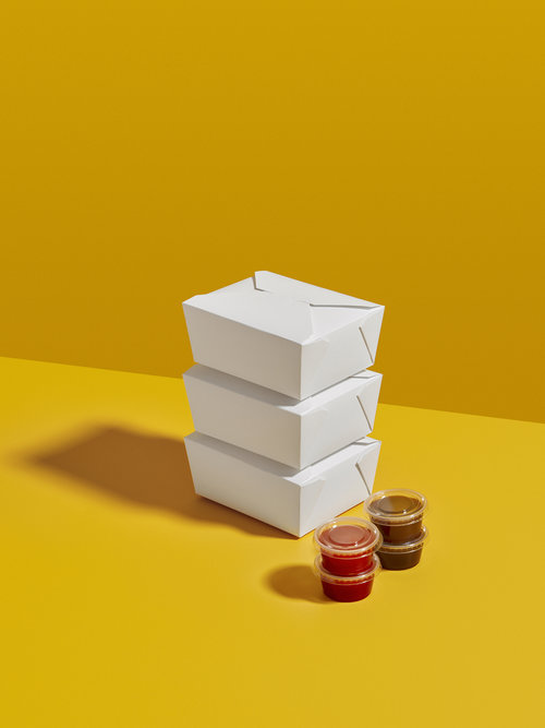 Three white takeout containers stacked on a yellow surface next to small sauce containers, creating a vibrant and simple studio shot.