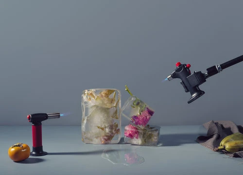 Still life photo of melting ice cubes containing flowers next to a persimmon, dishtowel, banana, and blow torches set against grey backdrop.