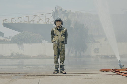 Full-length shot of a firefighter in uniform and helmet standing outdoors, with a water spray and industrial structure in the background.