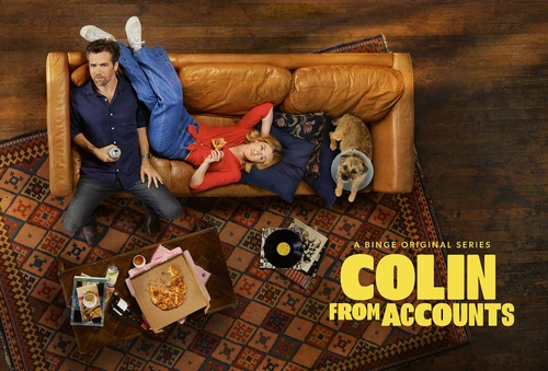 Promotional shot from above of the television show Colin From Accounts, featuring two actors, a dog, pizza, vinyl records, and furniture.