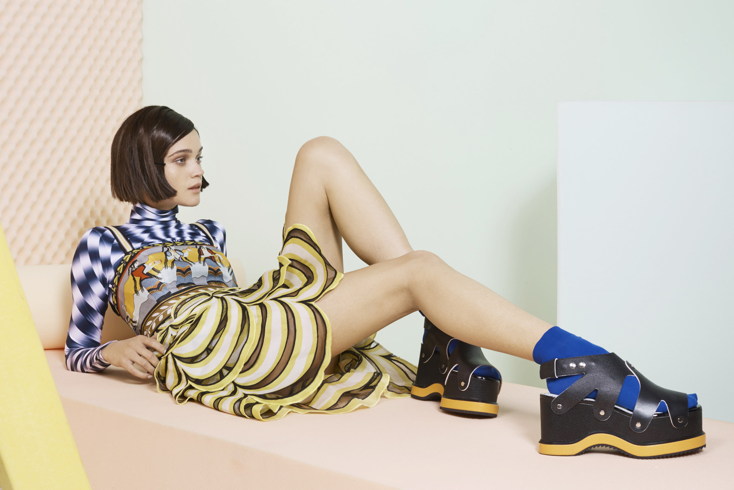 A brunette woman poses lying down in a patterned dress and black platform shoes against a split pastel blue and pink background.