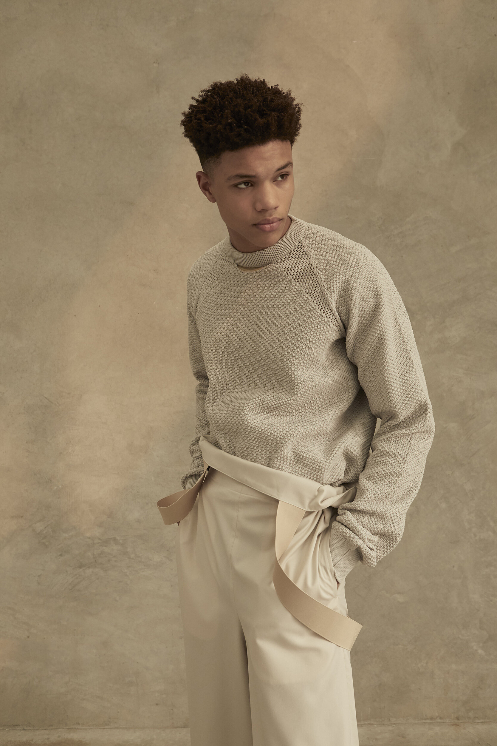 Young adult male fashion model in casual knit sweater and wide leg dress pants posing indoors against neutral mottled background.