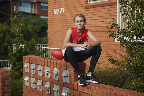 Young girl in a sports uniform seated on a brick wall outdoors with a sports ball next to a brick building and green foliage.