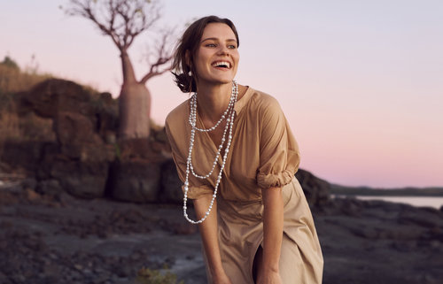 A smiling woman with pearl necklaces in a tan dress stands outdoors near a tree on a rocky surface against a pastel sky at dusk.