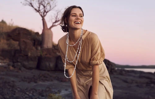 A smiling woman with pearl necklaces in a tan dress stands outdoors near a tree on a rocky surface against a pastel sky at dusk.