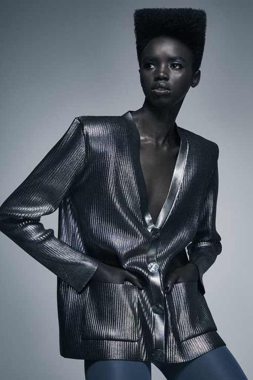 Stylish portrait of an adult woman with hands in pockets, wearing a metallic silver blazer and blue tights against a grey studio backdrop.