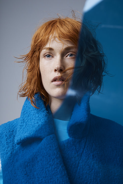 Close-up of a woman with red hair and blue eyes, partially obscured by a blue tinted glass or plastic pane, wearing a blue coat.