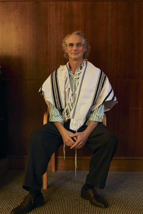 An adult man with curly gray hair is sitting on a chair indoors, wearing a green shirt, dark pants, dark shoes, and a white prayer shawl.