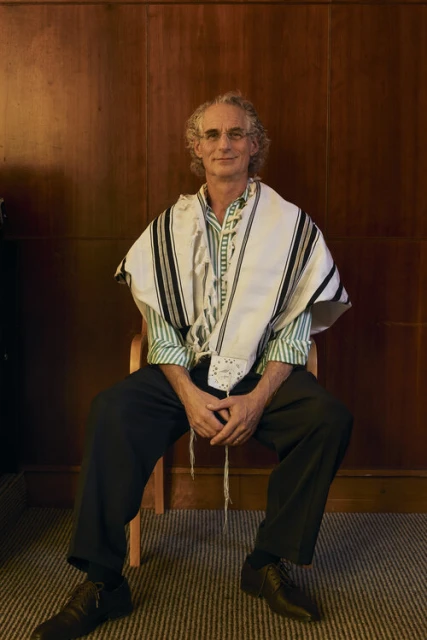 An adult man with curly gray hair is sitting on a chair indoors, wearing a green shirt, dark pants, dark shoes, and a white prayer shawl.
