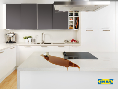 Modern kitchen interior featuring a spilled cup of brown liquid on a white countertop with white and gray cabinets, and IKEA logo.
