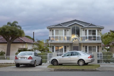 Two silver sedans parked in the driveway of a multi-story suburban house under an overcast sky.