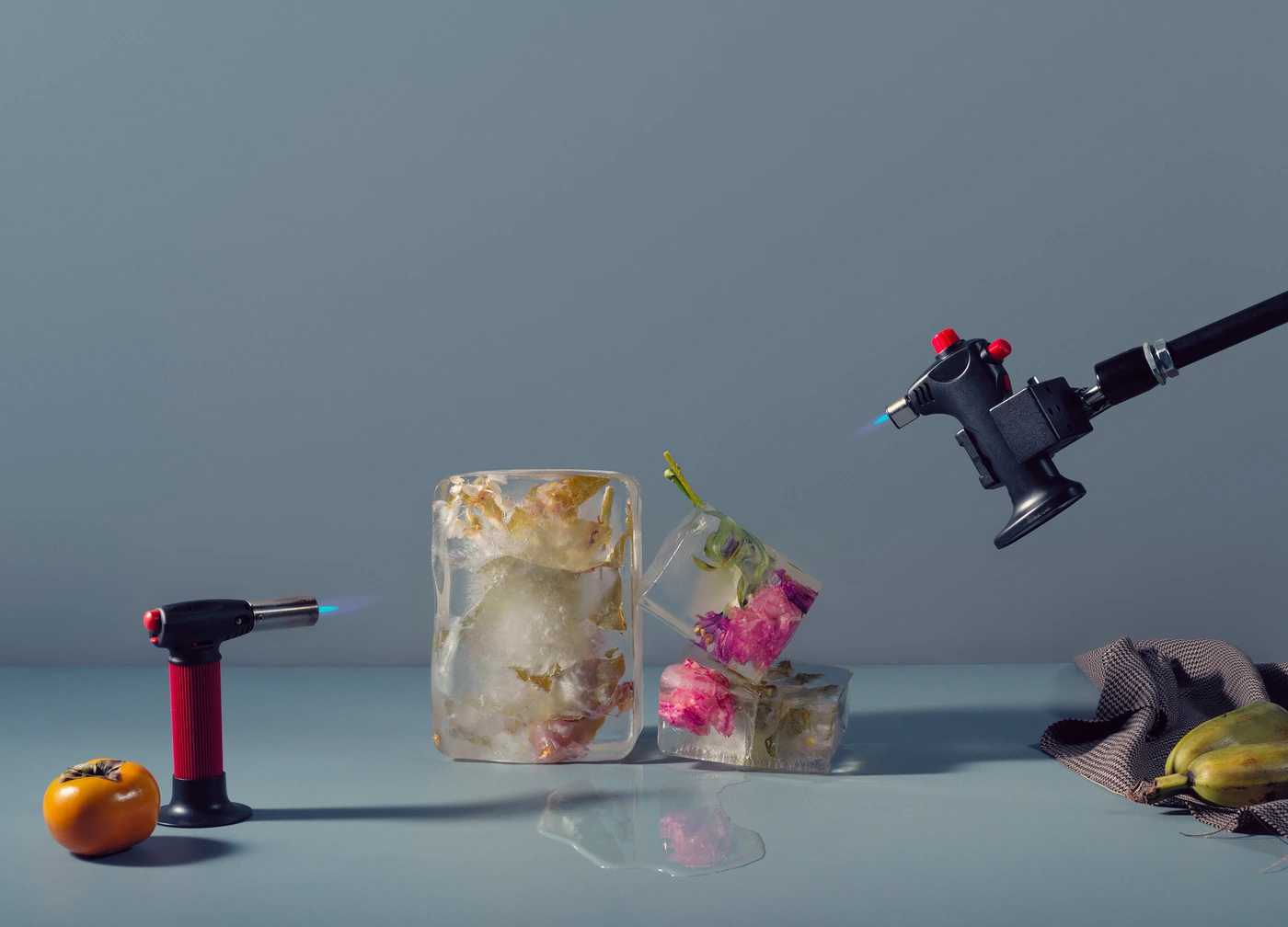 Frozen flowers inside ice blocks being melted by two handheld blowtorches on a grey surface with a persimmon and bananas.