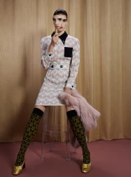 Model seated on a clear stool holding a hand mirror up to her face, wearing a patterned dress, knee socks, and gold shoes.