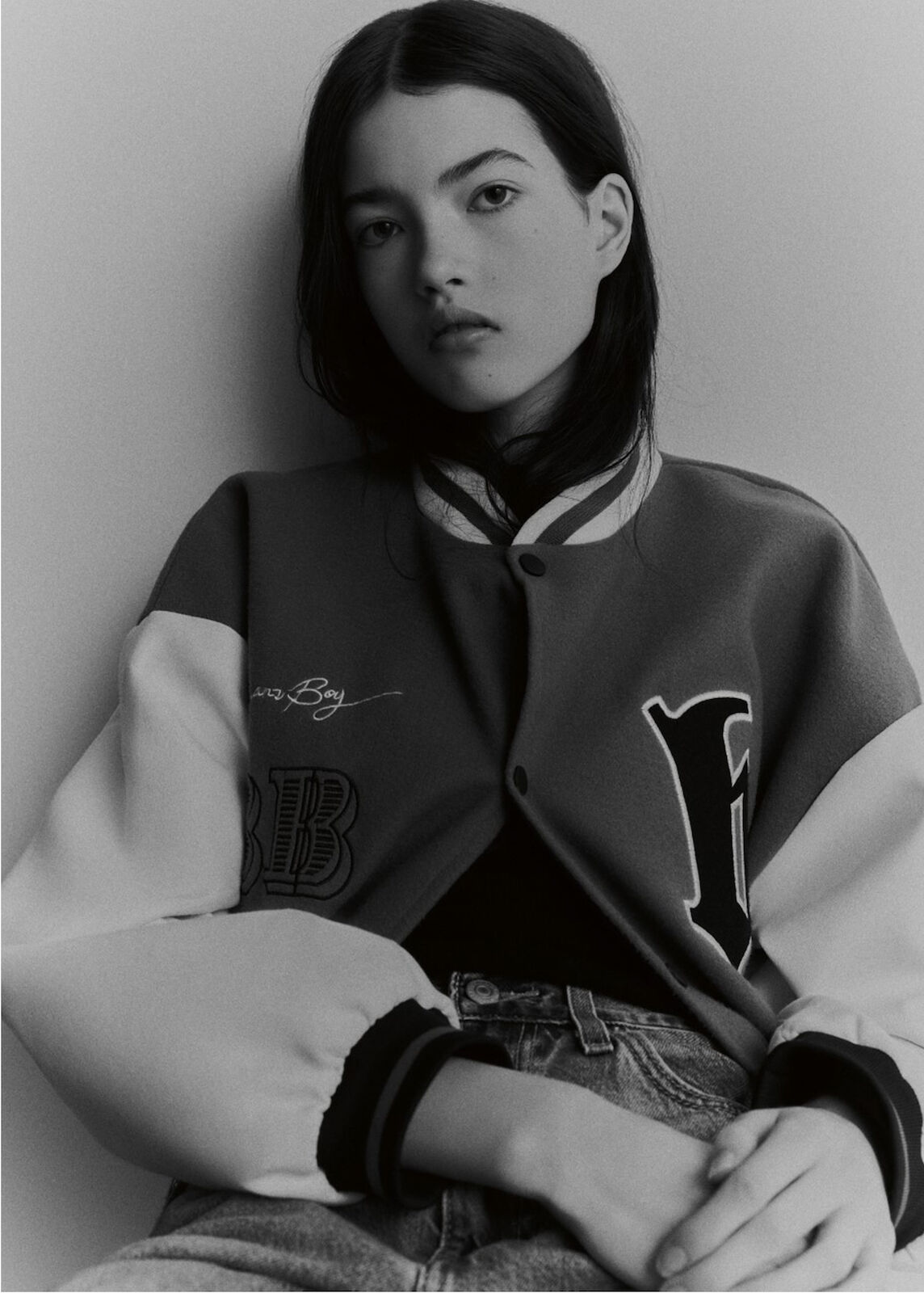 Portrait of a young woman in a red and white varsity jacket against a grey background, photographed indoors.