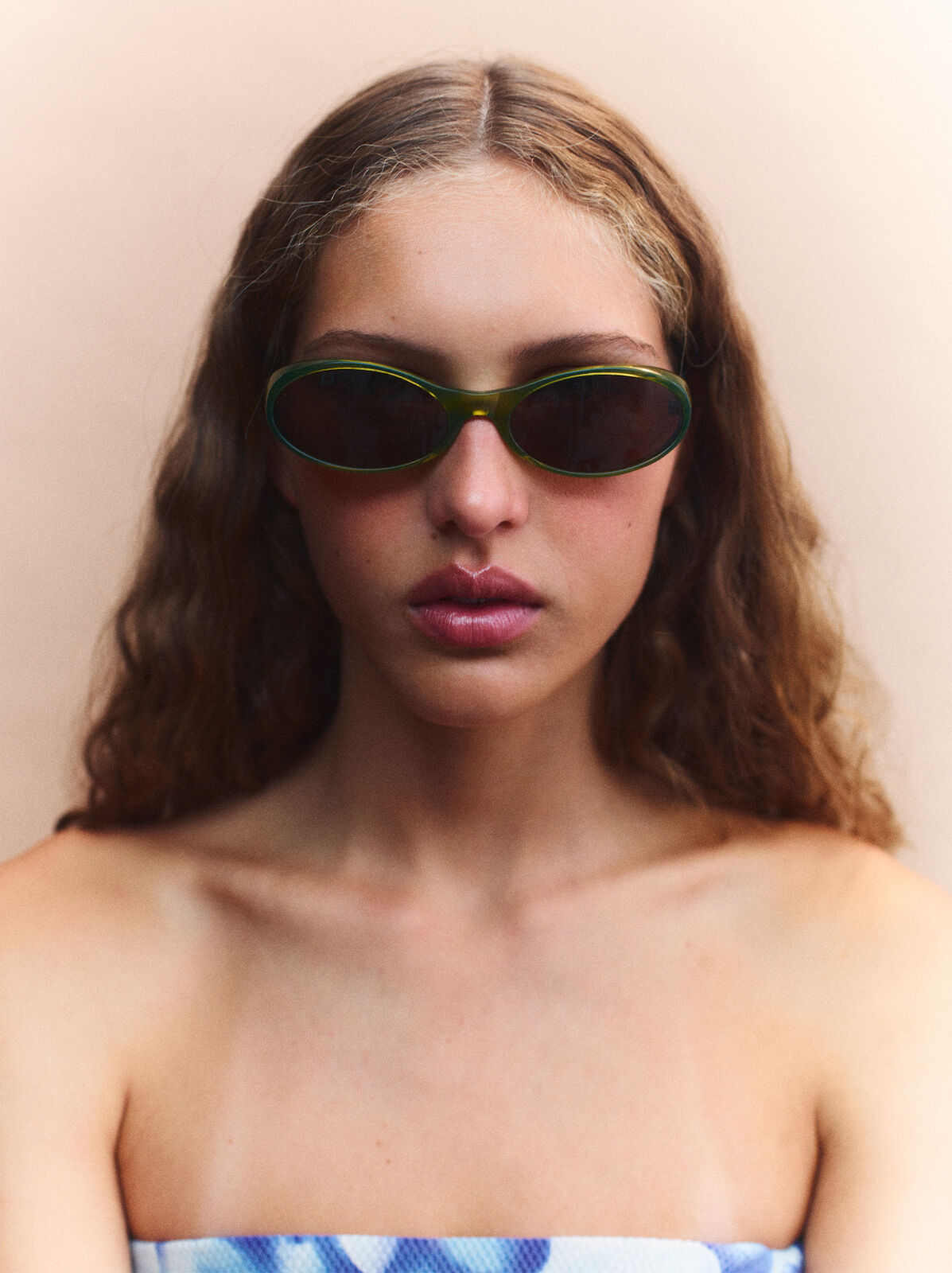 Close-up of a young woman with wavy brown hair and olive-framed sunglasses, wearing a strapless blue and white top against a light background.