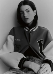 Close-up portrait of a young woman wearing a varsity jacket and denim jeans against a plain neutral background.