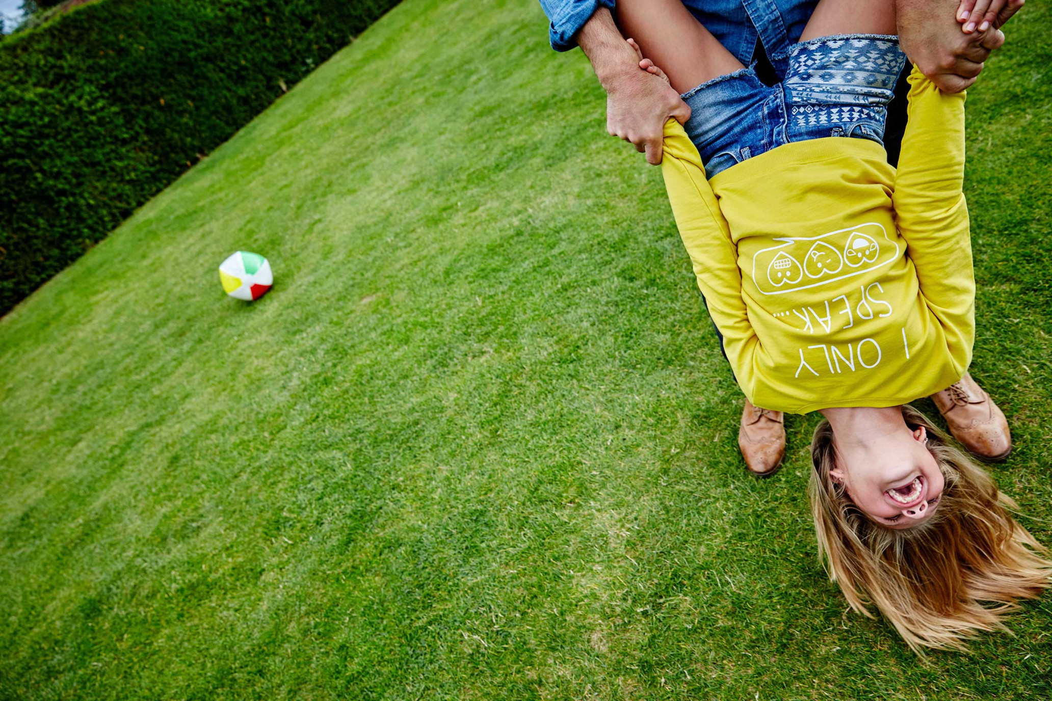 Child is playfully held upside down by an adult outdoors on green grass with a beach ball nearby, showcasing a fun and carefree moment.