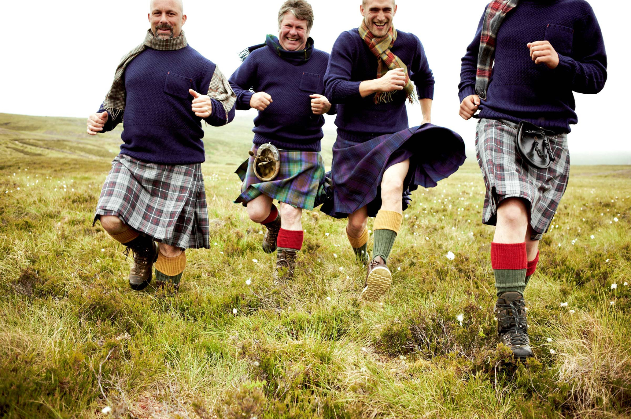 Four adult males in kilts run across a grassy field, wearing sweaters and scarves, showcasing Scottish traditional attire in a bright outdoor scene.