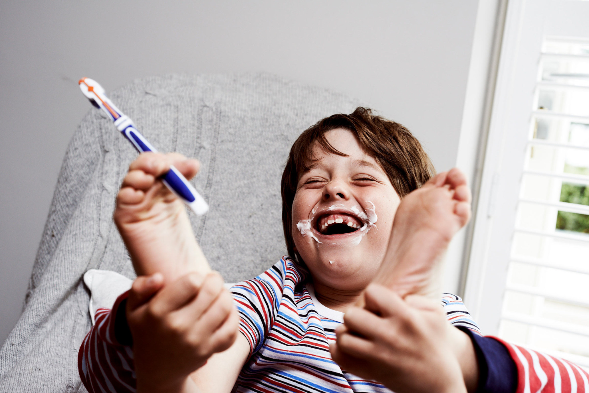 Laughing child holds feet, smeared with white cream, alongside a toothbrush in an indoor setting with filtered window light.