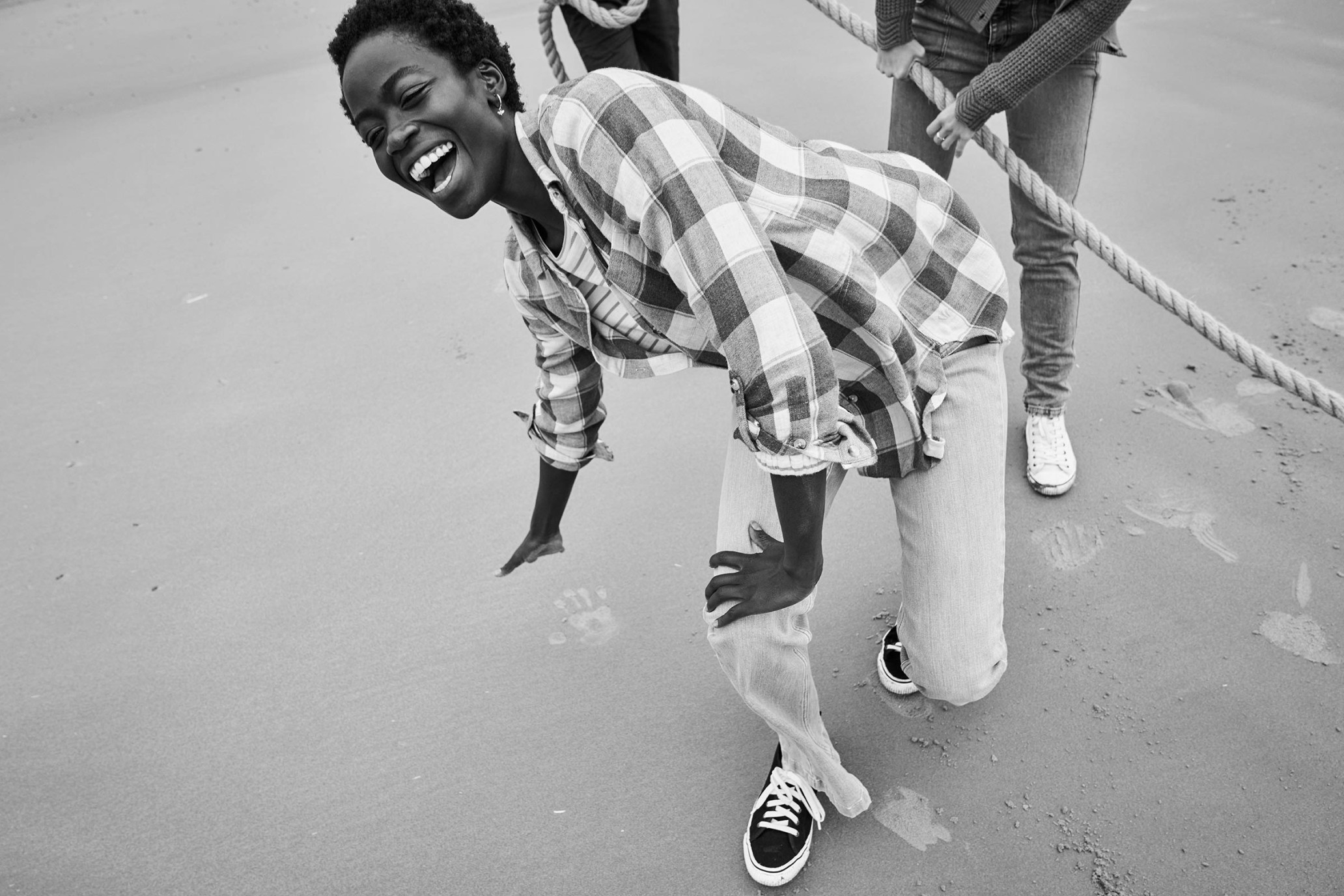 Laughing woman in checkered shirt and jeans, barefoot on a beach, pulling a rope with friends in a black and white photo.