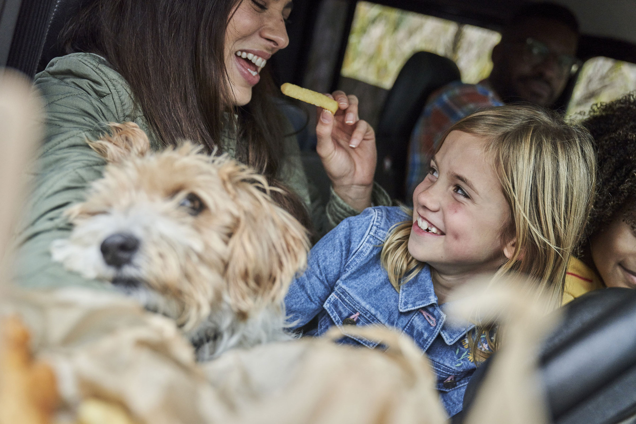 Smiling family of four with a dog inside a car on a road trip, the mother offering the child a snack with trees in the background.