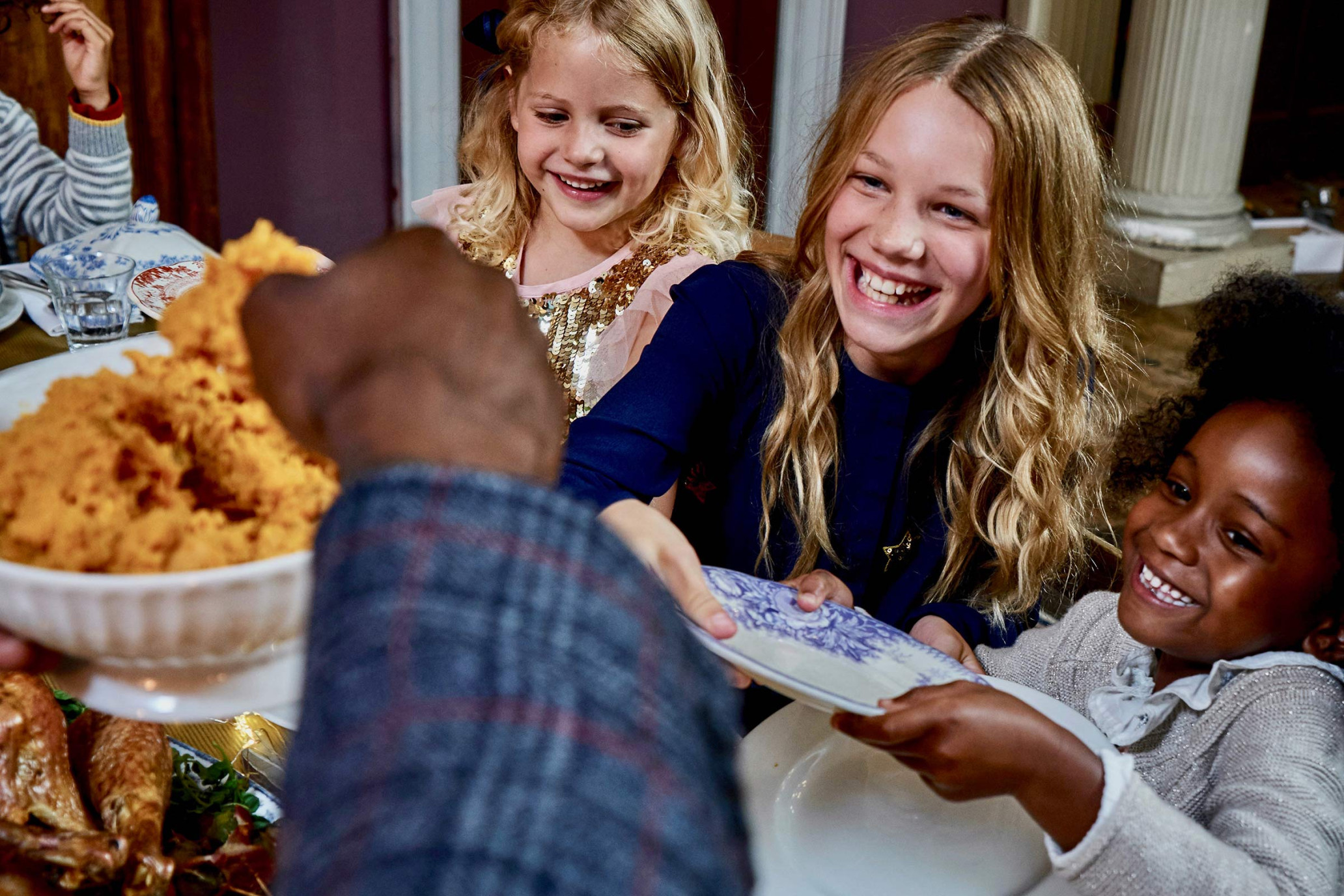 Three smiling girls at a dinner table reach for a plate being offered by a person in a plaid shirt, with holiday dishes visible.