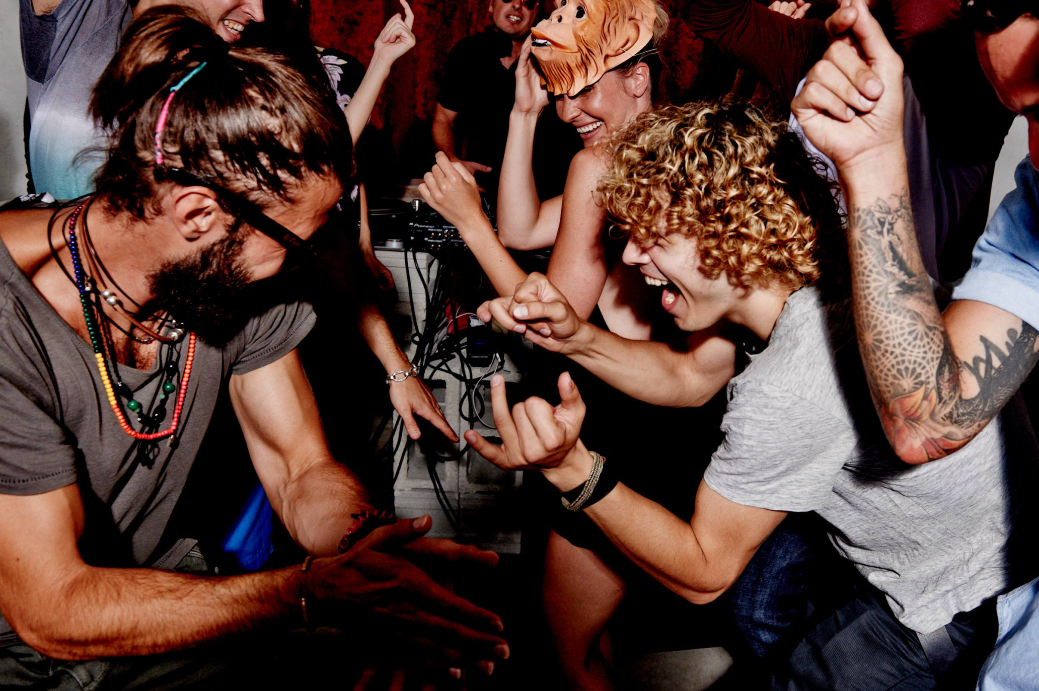 Excited group of young adults dancing at an indoor party or rave, with colorful lights, DJ equipment, and a person wearing a mask.