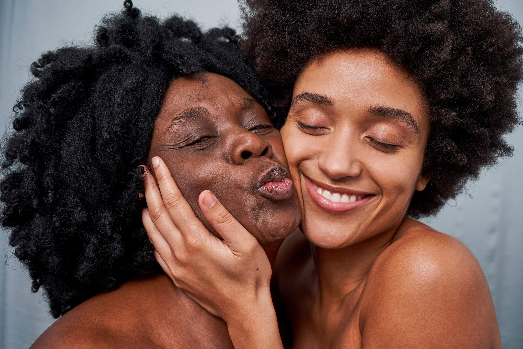 Close-up of two adult women with dark curly hair embracing indoors against gray wall, one puckering lips and the other smiling.