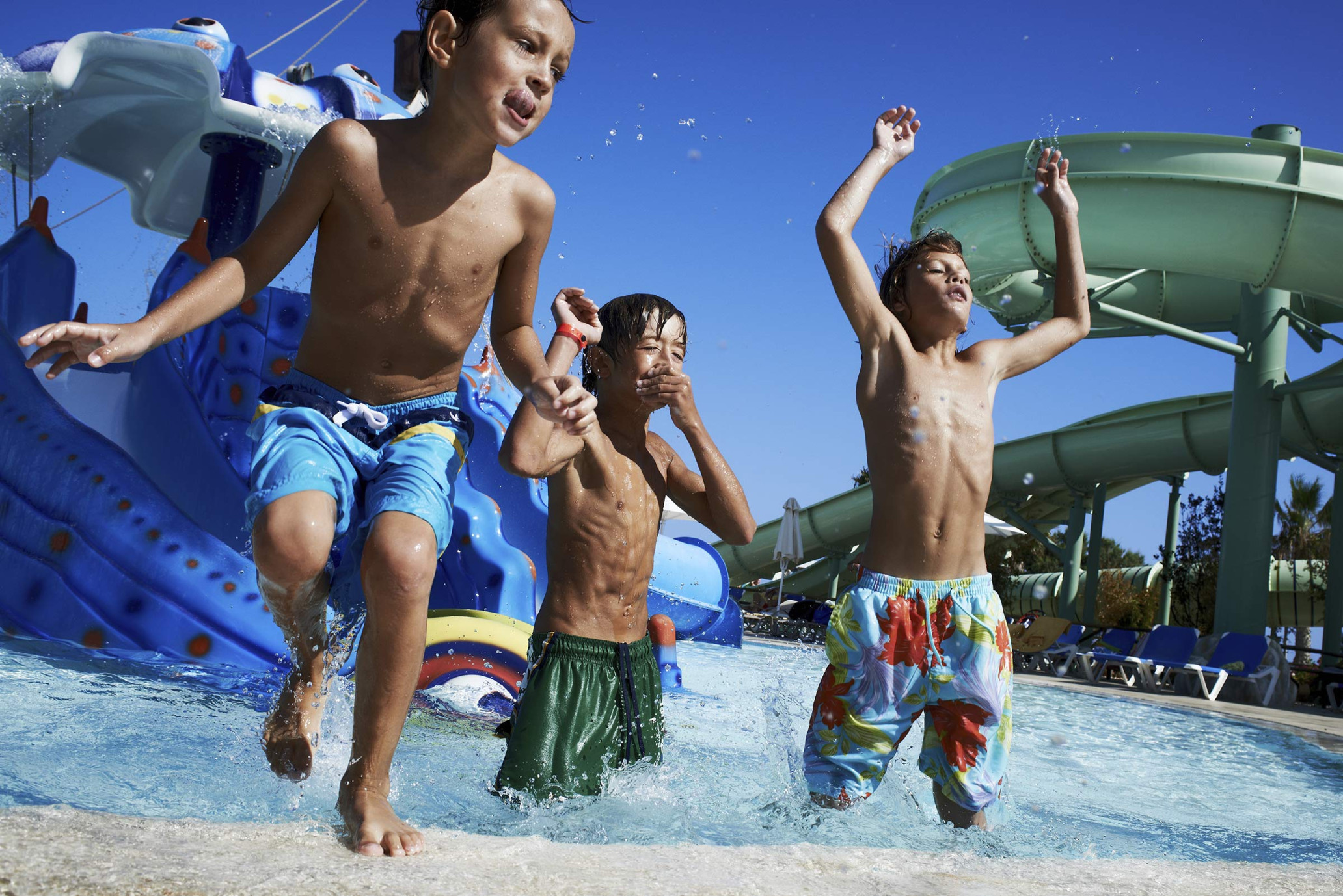 Three smiling children jumping in a pool with water park features under a bright blue sky, capturing a fun, sunny day at a water park.