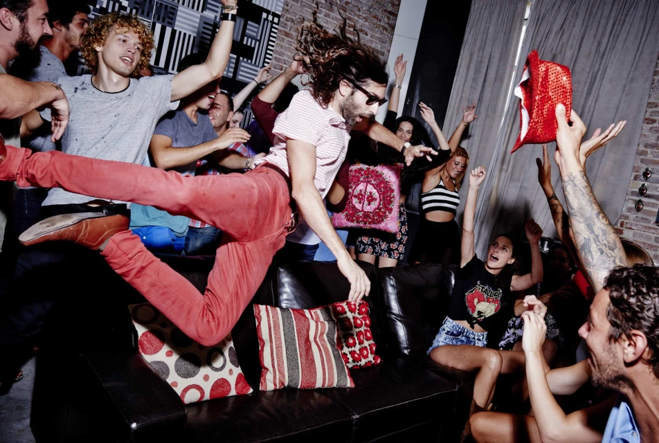 Enthusiastic group of young adults celebrating at an indoor party, one man jumping over a black sofa. Festive atmosphere, casual fashion, indoor setting.